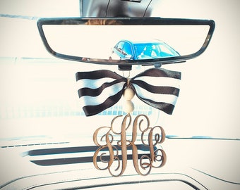 May include: A black and white striped bow with a wooden monogram charm hanging from the rearview mirror of a car. The monogram is the letters "KB" in a cursive font. The car's radio display shows the station "93.7 WHEL" and the artist "Kane Brown".