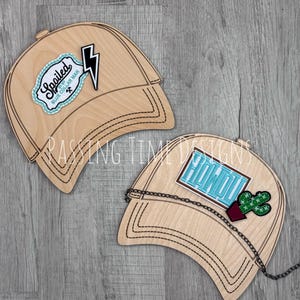 May include: Two wooden baseball cap cutouts with decorative patches. One cap has a lightning bolt and a patch that says "Spoiled Blue Collar Man". The other cap has a cactus and a patch that says "Howdy". Both caps have a chain.
