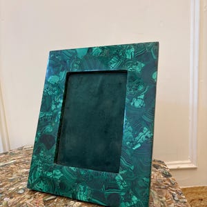 Puede incluir: Un marco de fotos rectangular con un diseño de malaquita. El marco es de color verde vibrante con patrones verdes oscuros y negros. El interior del marco es de color verde oscuro. El marco se muestra sobre una superficie decorativa.