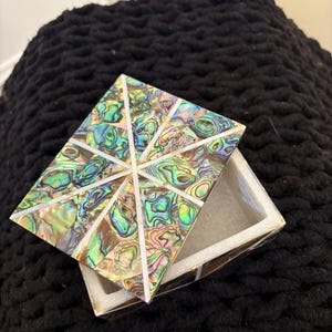 May include: A small, rectangular box with a hinged lid. The lid is made of iridescent mother-of-pearl tiles in shades of green, blue, and brown. The box is made of white marble and has a geometric design.
