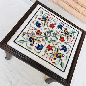 Puede incluir: Una mesa cuadrada de mármol blanco con un diseño floral incrustado con piedras de colores. La mesa tiene un marco de madera oscura y cuatro patas. La mesa presenta un diseño floral con pájaros y flores en tonos de azul, rojo, verde y amarillo.
