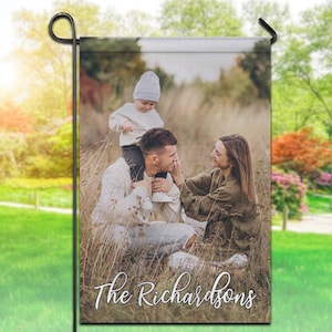 May include: A garden flag displaying a family portrait. The image shows a family of three in a field of tall grass. The text "The Richardsons" is printed at the bottom in a cursive font. The flag is designed for outdoor use.