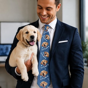 May include: A blue necktie featuring a repeating pattern of a golden retriever puppy face, white paw prints, white bone icons, and the name Rocky written in white script. The tie is worn with a white dress shirt and a dark navy blue suit jacket.