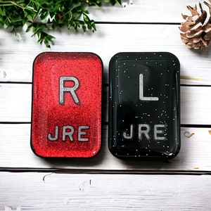 May include: Two small, square, glittery tiles with the letters "R" and "L" on them. The "R" tile is red with silver glitter and the "L" tile is black with silver glitter. Both tiles have the text "JRE" printed below the letter.