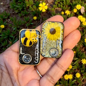 May include: Two rectangular, black and gold resin pendants. One features a yellow bee, the other a sunflower. Both have the letters "LARS" and a circular element. The background includes green plants and yellow flowers.