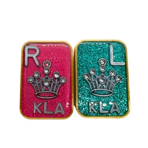 May include: Two square tiles with a gold border and glitter background. The tile on the left is pink with the letter "R" and the word "KLA" printed on it. The tile on the right is teal with the letter "L" and the word "KLA" printed on it. Both tiles feature a silver crown with rhinestones.