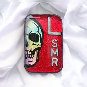 May include: A rectangular, resin-covered object with a red glitter background. A colorful skull graphic is on the left, and the letters "LSMR" are on the right. The object has a black border and is set against a white fabric.