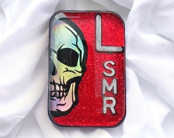 May include: A rectangular, resin-covered object with a red glitter background. A colorful skull graphic is on the left, and the letters "LSMR" are on the right. The object has a black border and is set against a white fabric.