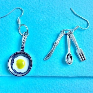 May include: A pair of silver earrings. One earring is a frying pan with a fried egg inside. The other earring is a knife, spoon, and fork.