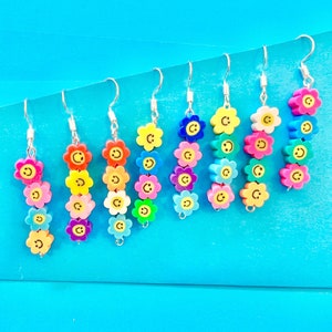 May include: A set of eight pairs of dangle earrings featuring colourful flower charms with smiley faces. Each earring has five flower charms in a variety of colours, including yellow, orange, pink, purple, blue, and green.