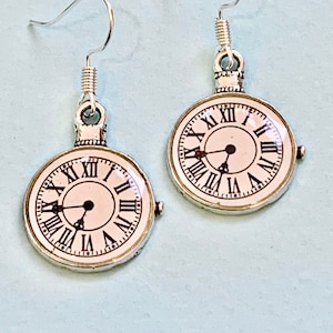 May include: A pair of silver dangle earrings with a white clock face featuring Roman numerals. The clock face is surrounded by a silver frame.