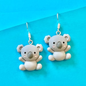 Op de afbeelding: Een paar zilveren oorhangers met grijze koalabeerbedels. De koala's hebben zwarte ogen, neuzen en monden en roze wangen.