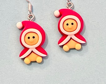 May include: A pair of red and white Christmas-themed earrings featuring a small, cartoon-like figure wearing a Santa hat. The figure has a yellow face with black eyes and a small, red mouth.