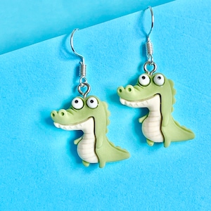 May include: A pair of green alligator earrings with white eyes and a silver hook. The earrings are on a blue background.