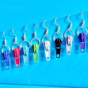 May include: Eight pairs of zipper earrings in various colors, including purple, blue, green, white, black, and red. The earrings are hanging from silver hooks.
