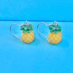 May include: A pair of silver dangle earrings with a yellow pineapple charm. The pineapple has a green top and a textured surface.