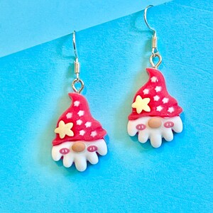 May include: A pair of silver dangle earrings featuring a red and white gnome design. The gnome has a white beard, a red hat with white stars, and a yellow star on top of the hat.
