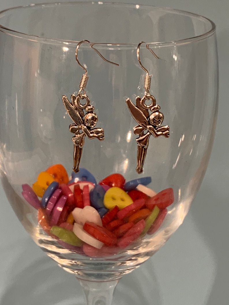 Puede incluir: Un par de pendientes de plata con un dise&ntilde;o de hada. Las hadas sostienen una peque&ntilde;a bola en sus manos. Los pendientes cuelgan de una copa de vino de vidrio.