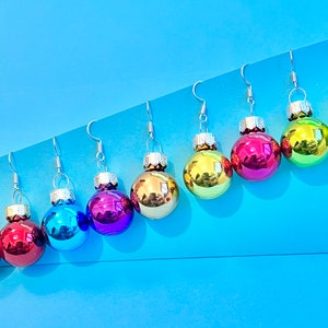 May include: Eight pairs of dangle earrings featuring small, round, colorful ornaments that resemble Christmas tree ornaments. The ornaments are in shades of red, green, blue, gold, and purple.