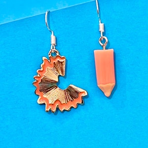 Könnte beinhalten: Ein Paar goldene und pfirsichfarbene Ohrringe in Form eines Bleistifts und eines Bleistift-Spans. Die Ohrringe hängen an silbernen Haken.