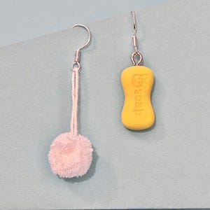 May include: A pair of quirky earrings. One earring features a small, fluffy, white pom-pom. The other earring has a yellow, soap-shaped charm with the word "soap" printed on it. Both earrings have silver-colored ear wires.