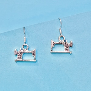 May include: A pair of silver earrings shaped like vintage sewing machines. The earrings are hanging from silver hooks.