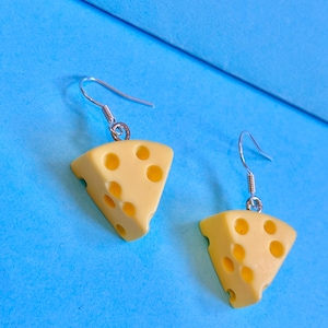 Könnte beinhalten: Ein Paar ausgefallene Ohrringe mit keilförmigen Käsescheiben mit Löchern. Die Ohrringe sind gelb mit silberfarbenen Ohrhaken, vor einem hellblauen Hintergrund. Ein lustiges und skurriles Accessoire.