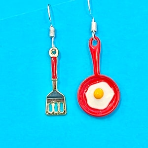May include: A pair of red and gold earrings featuring a frying pan with a fried egg and a spatula.