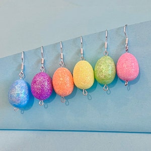 May include: Six pairs of dangle earrings with silver hooks. Each earring features a glittery oval-shaped charm in a different pastel colour: light blue, purple, orange, yellow, green, and pink.