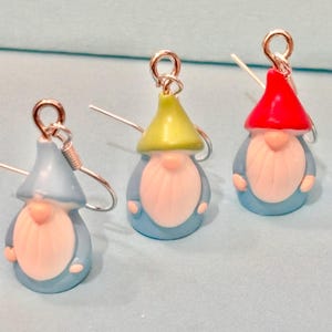 Può includere: Tre orecchini a forma di gnomo con ganci argentati. Ogni gnomo ha un corpo blu, una barba bianca e un cappello colorato: blu, verde e rosso. Gli orecchini sono posti su uno sfondo azzurro.