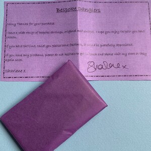 Puede incluir: Una tarjeta de agradecimiento morada con el texto "Bespoke Danglers" escrito en ella. La nota agradece al cliente su compra y menciona una amplia gama de pendientes hechos a medida. La nota tambi&eacute;n anima al cliente a dejar comentarios y visitar la tienda en Etsy.