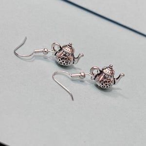 Peut inclure: Une paire de boucles d'oreilles en forme de théière argentée avec un design détaillé. Les boucles d'oreilles sont sur un fond bleu clair.