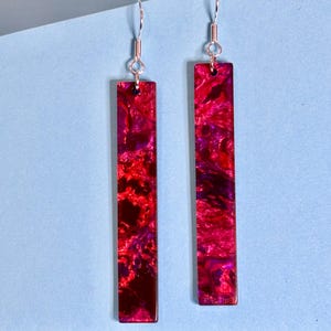 May include: A pair of rectangular earrings with a marbled pattern in shades of red and purple. The earrings hang from silver-coloured hooks. The background is a light blue.