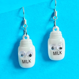 May include: A pair of white milk carton earrings with googly eyes and the word "MILK" printed on them. The earrings are hanging from silver hooks.