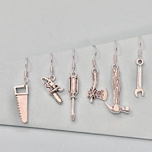 May include: Silver-toned tool-themed earrings. The set includes earrings shaped like a saw, chainsaw, screwdriver, axe, hammer, and wrench. Each earring hangs from a simple hook, showcasing the detailed miniature tool designs.