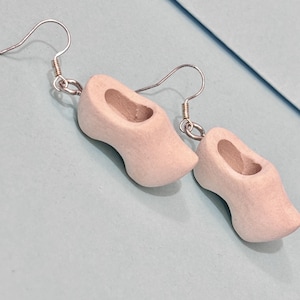 Puede incluir: Un par de pendientes de madera con forma de zueco de color beige claro. Los pendientes tienen ganchos plateados.