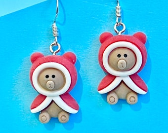 May include: A pair of silver dangle earrings featuring a red and white teddy bear wearing a red hooded coat. The teddy bears have brown eyes and noses.