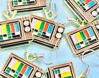 May include: Six pairs of gold-tone earrings shaped like vintage televisions with a colourful static screen pattern. Each earring has a silver hook.