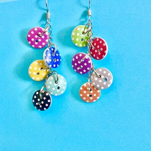 Puede incluir: Un par de pendientes colgantes con una cadena plateada y varios botones de lunares de colores. Los botones son en tonos de rosa, verde, azul, amarillo, naranja y gris.