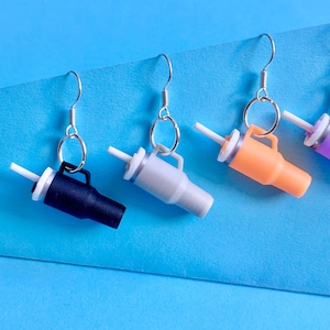 Puede incluir: Cuatro pares de pendientes colgantes con vasos de plástico en miniatura y pajitas. Los vasos son de color negro, gris, naranja y morado.