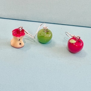 May include: Three pairs of apple-shaped earrings. One pair is a red apple, one pair is a green apple, and one pair is a bitten apple with a white core.