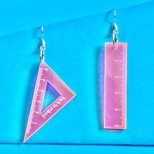 May include: Pink plastic earrings shaped like a ruler and a triangle. The triangle has the text "Back to School" printed on it.