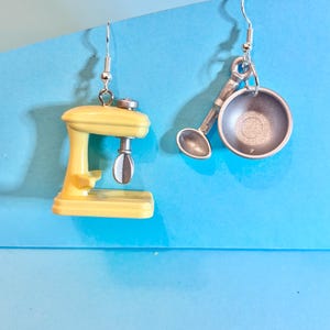 Può includere: Un paio di orecchini a forma di utensili da cucina in miniatura. Un orecchino è un mixer giallo, l'altro una padella e un cucchiaio argentati. Entrambi gli orecchini hanno chiusure a gancio argentate, su sfondo blu.