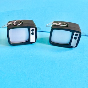 Puede incluir: Un par de pendientes de televisión en blanco y negro. Cada pendiente presenta un pequeño televisor de estilo retro con una pantalla blanca y un marco negro. Los pendientes tienen ganchos plateados.
