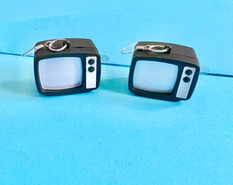 May include: A pair of black and white television earrings. Each earring features a small, retro-style TV with a white screen and a black frame. The earrings have silver-coloured ear wires.