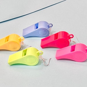 Op de afbeelding: Vijf kleurrijke plastic fluitjes oorbellen. De oorbellen zijn in de kleuren blauw, geel, groen, roze en rood. Elk fluitje heeft een zilveren haak.