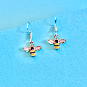 May include: A pair of silver bee earrings with yellow and black stripes and white wings. The earrings are on a light blue background.