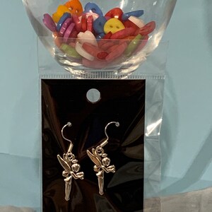 Puede incluir: Un par de pendientes de plata con un dise&ntilde;o de Campanilla. Los pendientes est&aacute;n empaquetados en una tarjeta negra con una bolsa de pl&aacute;stico transparente.