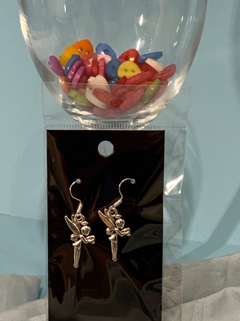 Puede incluir: Un par de pendientes de plata con un dise&ntilde;o de Campanilla. Los pendientes est&aacute;n empaquetados en una tarjeta negra con una bolsa de pl&aacute;stico transparente.