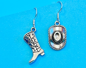 May include: Silver-toned cowboy boot and cowboy hat earrings. The boot features a detailed design with a blue accent, while the hat has a rope-like border. Both earrings hang from a simple silver hook, perfect for a Western-themed look.